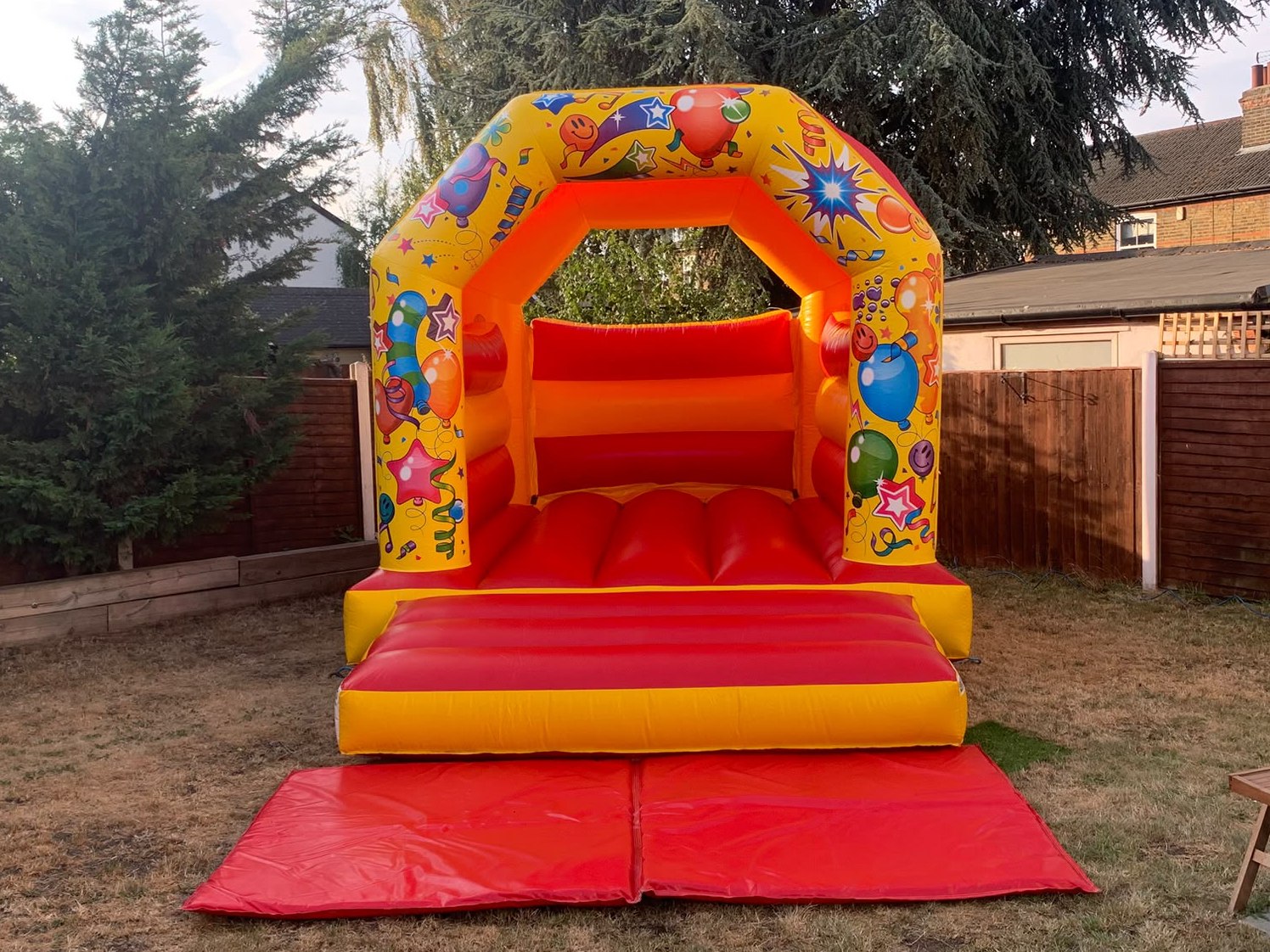 Red and yellow Tiny Tots bouncy castle with balloon artwork and front safety mat in a garden